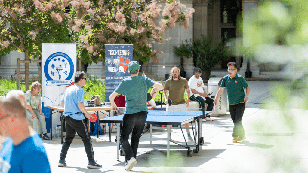 Unser Wiener Tischtennis Verein im Rathaus: Engagierte Mitglieder aus der Tischtennis-Community zeigen Besuchern an einer Outdoor-Tischtennisplatte Tipps und Tricks, während im Hintergrund der rege Informationsaustausch beim Jeder mit Jedem-Event stattfindet.
